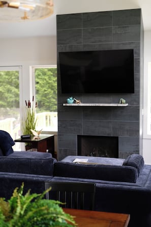 Ein modernes Wohnzimmer mit einem großen Flachbildfernseher an einer dunkel gefliesten Wand, einem Ecksofa und einem kleinen Kamin mit Dekorationselementen.