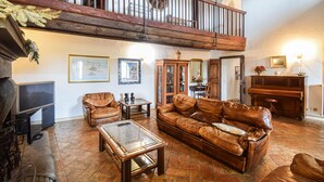 A spacious living room with a high ceiling, a wooden staircase, a fireplace, leather furniture, and a piano.