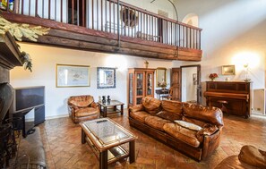 A spacious living room with a high ceiling, a wooden staircase, a fireplace, leather furniture, and a piano.
