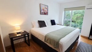 Ein Schlafzimmer mit Bett, Nachttisch, Lampe, Kunstwerk, Fenster und einem karierten Teppich.