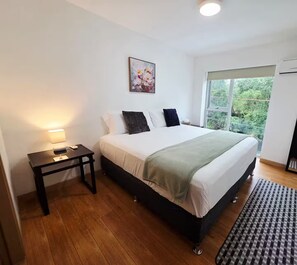 Ein Schlafzimmer mit Bett, Nachttisch, Lampe, Kunstwerk, Fenster und einem karierten Teppich.