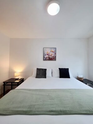 Een hotelkamer met een bed, nachtkastje, lamp, schilderij en een klein mandje.