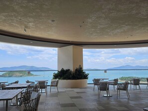 An outdoor seating area with tables and chairs, a large planter with greenery, and a view of the sea and mountains. - The Paramount Apartment Nha Trang (Bac Nha Trang)