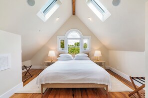 Ein Schlafzimmer mit schrägverdeckter Decke, Dachfenstern, einem Bett mit weißer Bettwäsche, zwei Nachttischlampen und Blick auf Grünfläche.