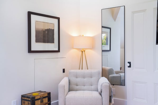 A room with a framed picture, a floor lamp, a mirror, a chair, and a small wooden chest.