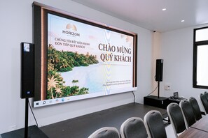 A conference room with a large screen displaying a presentation, a projector, a laptop, and rows of chairs. - HORIZON MINH CHAU HOTEL (Van Don)