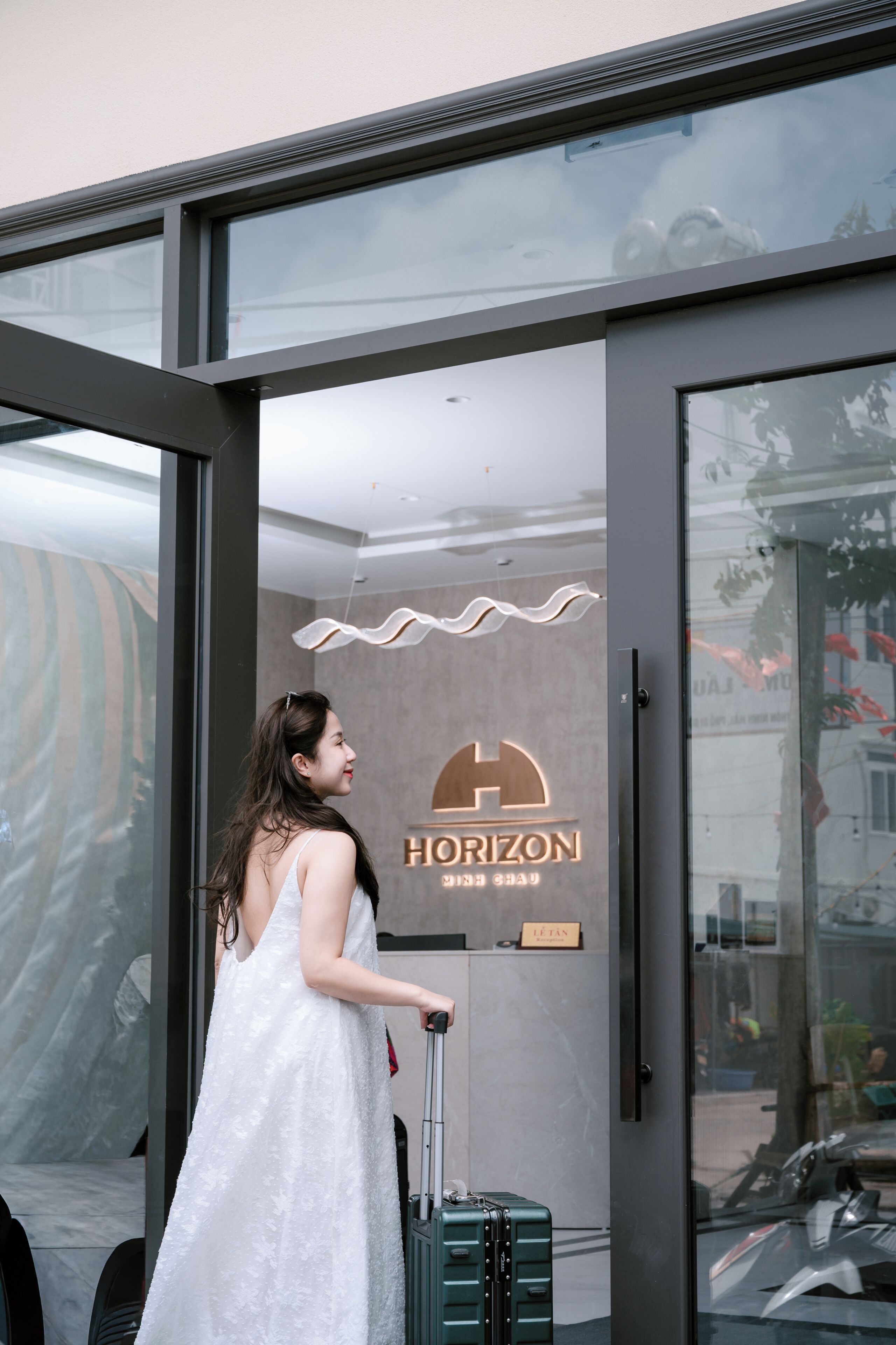 A woman in a white dress exiting a hotel with a suitcase, the hotel's name 'HORIZON' is visible on the door.