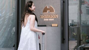 A woman in a white dress exiting a hotel with a suitcase, the hotel's name 'HORIZON' is visible on the door.