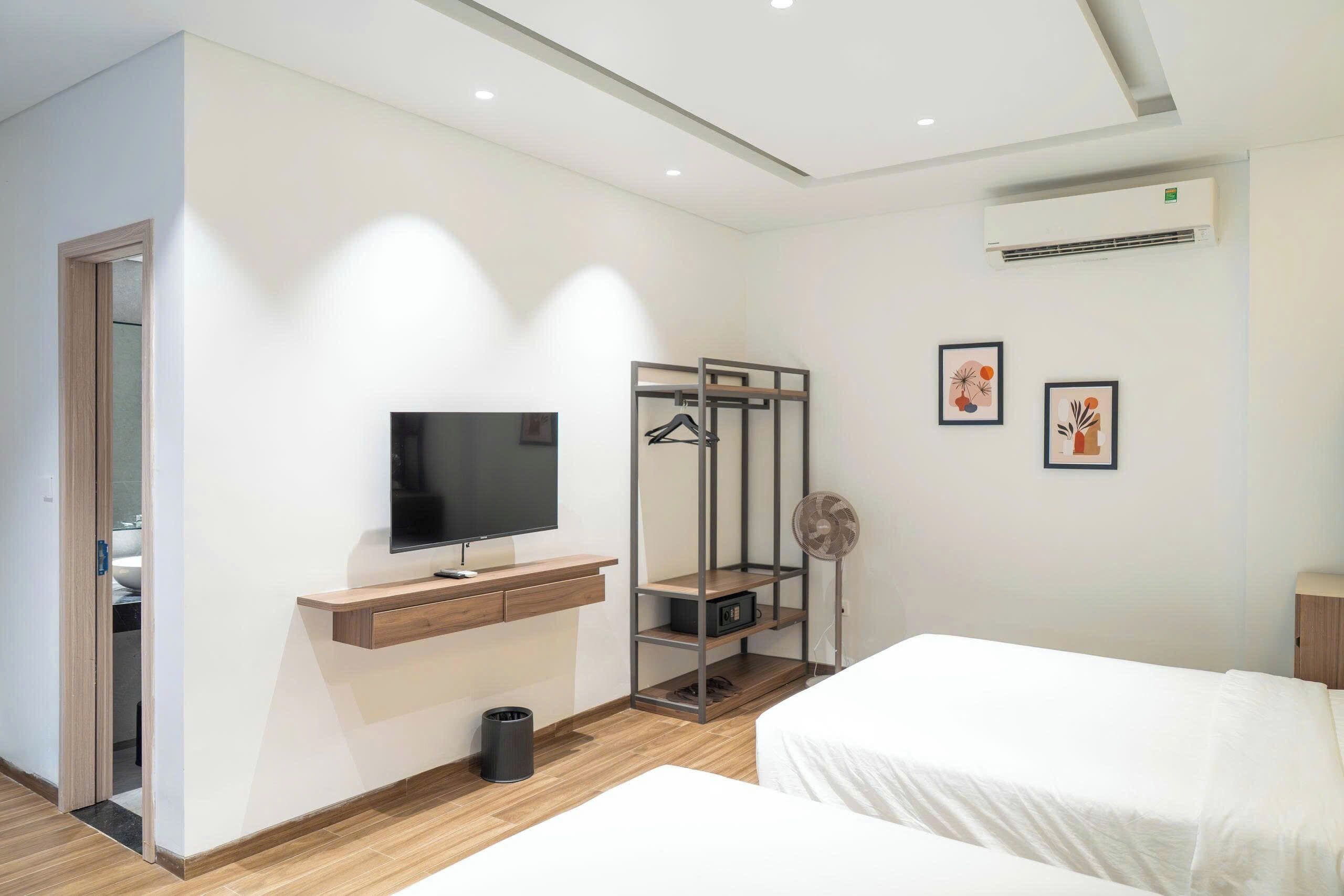 A hotel room with a flat-screen TV, a wooden desk, a hanging coat rack, a small fan, and two beds.