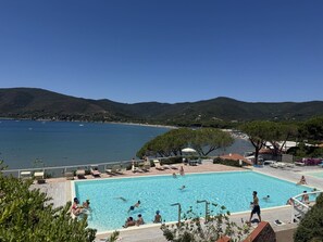 Ein Schwimmbecken mit Personen, umgeben von einer Sonnenterrasse und mit Blick auf ein Gewässer und Hügel.