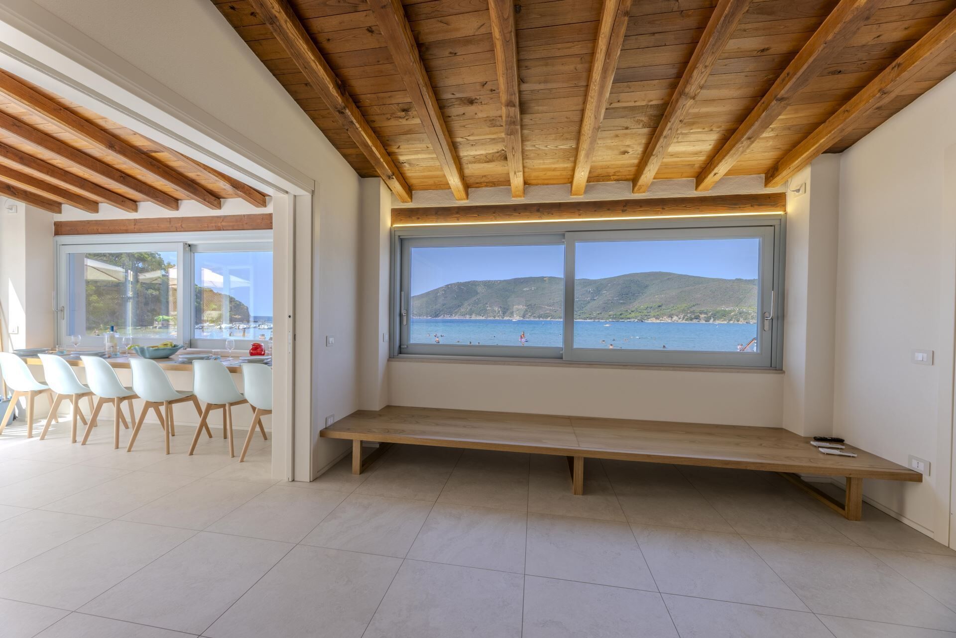 Ein modernes Interieur mit Holzbalkendecke, einem großen Fenster mit Blick auf ein Gewässer und einer Sitzecke mit einer Bank.