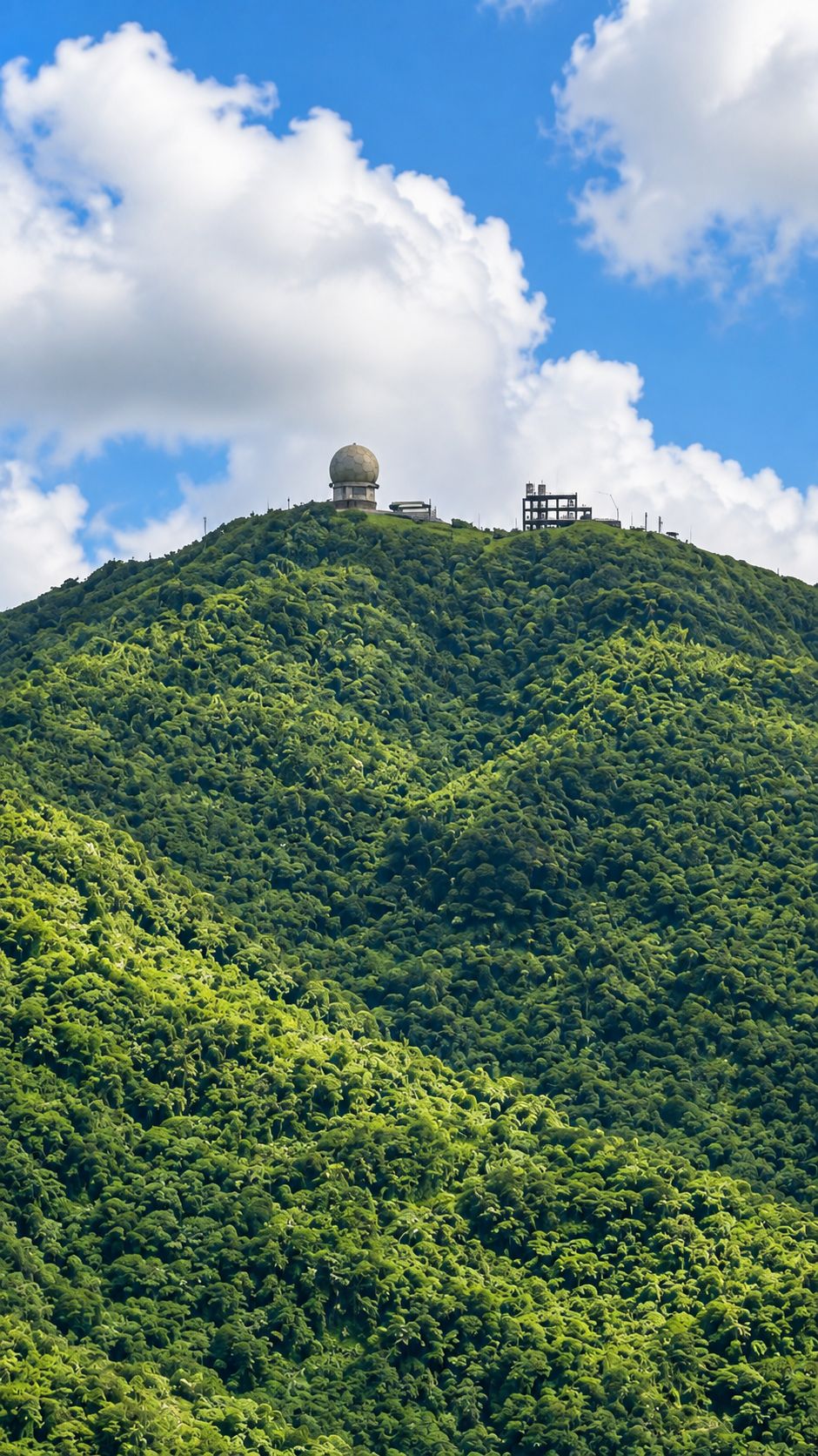 Une colline verdoyante et luxuriante, surmontée d’une structure sphérique, qui pourrait être une station météorologique, et d’un petit bâtiment doté d’un toit plat.