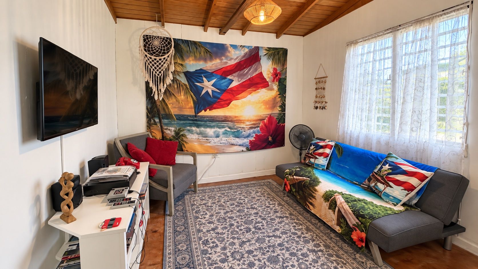 Een woonkamer met een televisie, een bureau met een computer, een bank met een vlaggedekbed, een groot schilderij van een strandgezicht en een kamerkleed met een patroon.