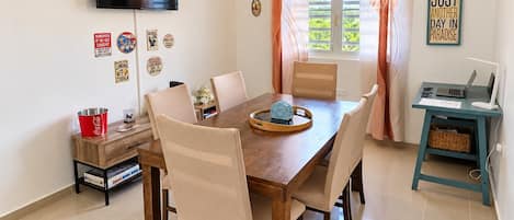 Un comedor con mesa de madera, sillas, televisor montado en la pared y ventana con cortinas.