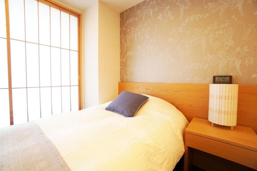 Quarto com cama, mesa de cabeceira, relógio de parede e janela com padrão de grade.