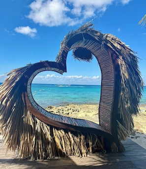 A heart-shaped structure made of palm leaves and metal, overlooking a beach with clear blue water and boats. - Rosa & Cioccolato (Cozumel)