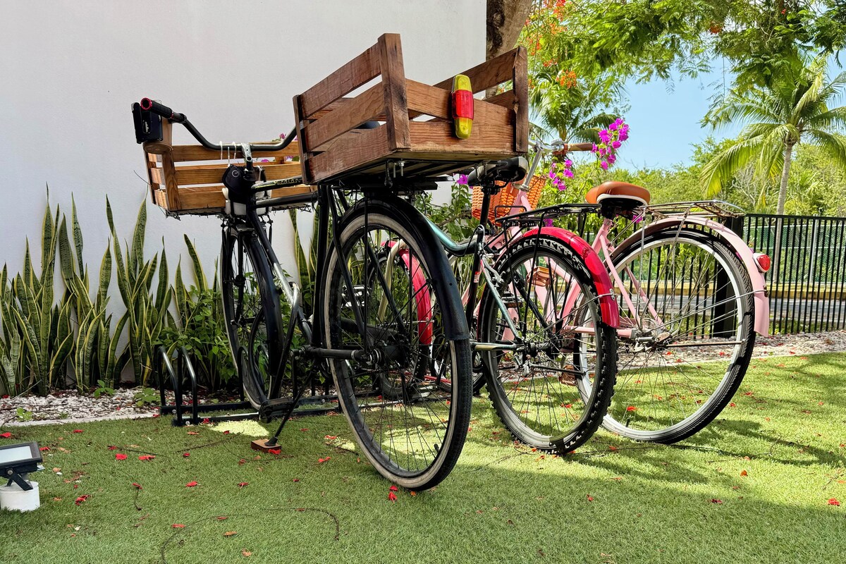 Two bicycles parked on a grassy area with a fence and plants in the background.