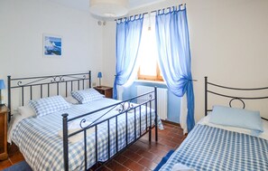 Ein Schlafzimmer mit einem Doppelbett, einem Einzelbett, einem Nachttisch, einer Lampe, einem Fenster mit blauen Vorhängen und einer Wandleuchte.