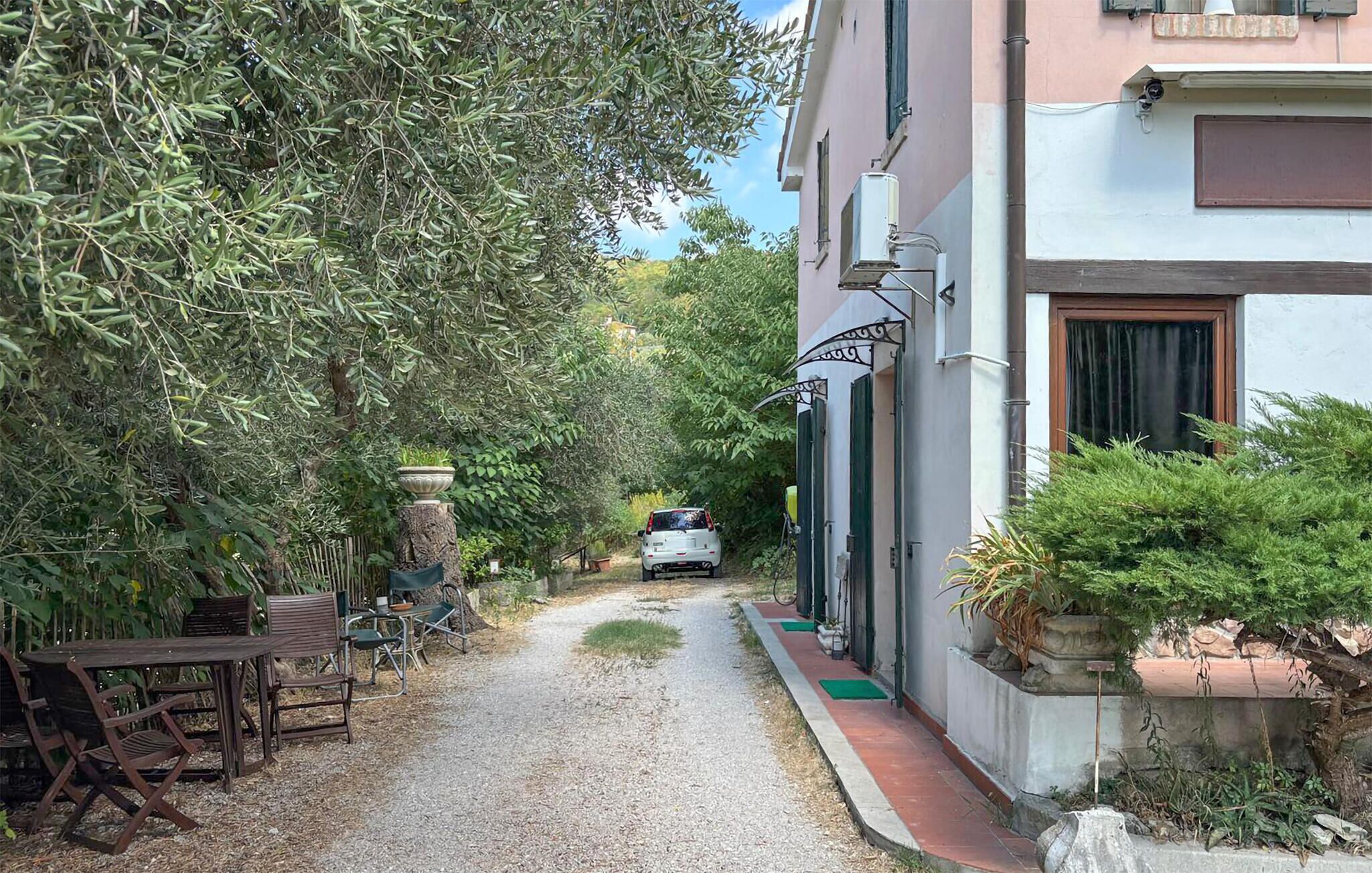 Une ruelle étroite avec un chemin de gravier, une voiture blanche garée au bout, et un bâtiment à la façade blanche avec une porte surmontée d’un portail en métal.