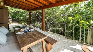 Un patio al aire libre con mesa de madera dispuesta para comer, rodeado de barandilla metálica y abundante vegetación.