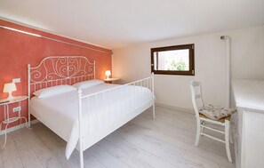 Ein Schlafzimmer mit weißem Metallbettgestell, einem Nachttisch mit Lampe, einem Sessel mit Kissen und einem Fenster mit Blick auf Grünflächen.