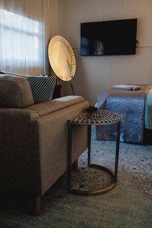 Ein Zimmer mit einer Couch, einem kleinen runden Tisch, einem Bett, einem an der Wand befestigten Fernseher und einem gemusterten Teppich.