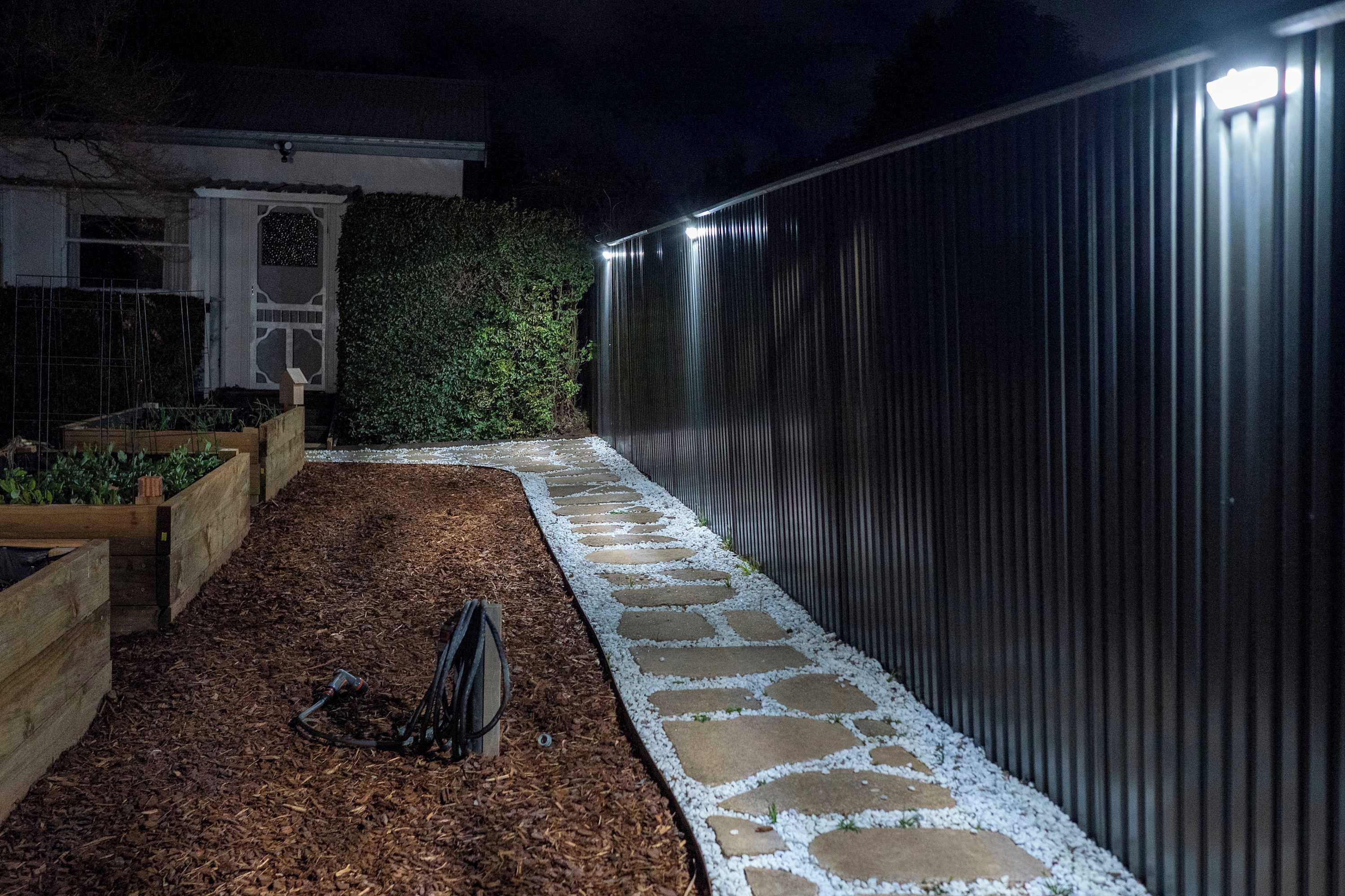 A garden pathway with a stone border, wooden raised beds, and a metal fence.