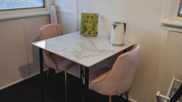 Un pequeño comedor con una mesa de mármol blanco, dos sillas rosas, un libro verde titulado 'THE GARDEN CHEFEN' y un hervidor blanco.