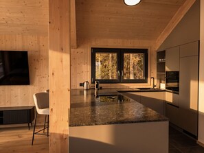 A modern kitchen with a central island, wooden walls, and a large window with a forest view. - 02_NTH055_18f_Sup Chalet mit Sauna und Hot Tub 2 E by Interhome (Großveitsch)