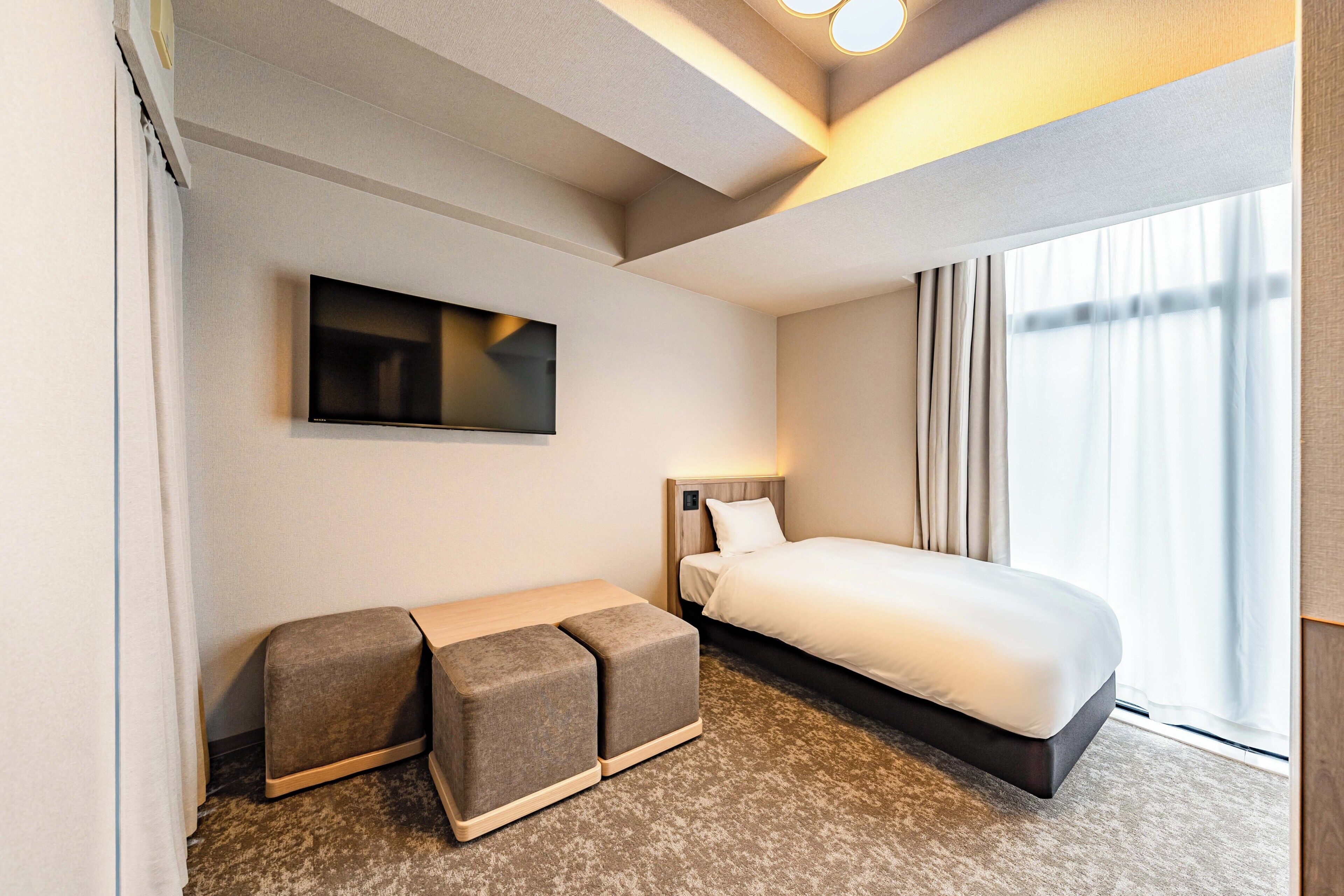Habitación de hotel con cama, televisor de pantalla plana en la pared, una pequeña zona de estar con otomanas y una ventana con cortinas.