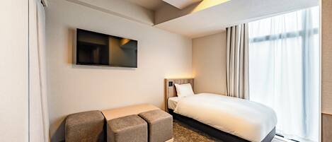 Habitación de hotel con cama, televisor de pantalla plana en la pared, una pequeña zona de estar con otomanas y una ventana con cortinas.