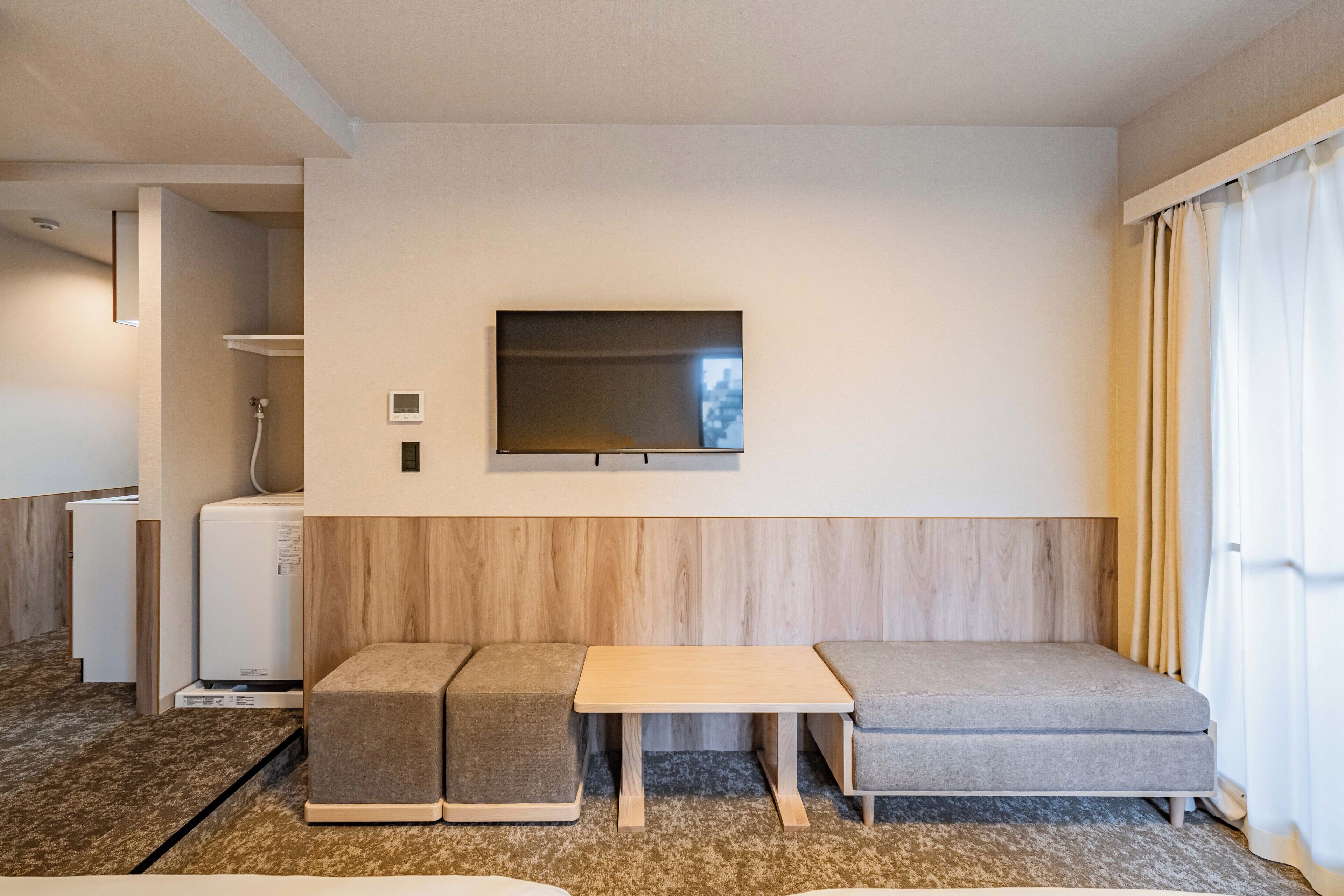 A hotel room with a sofa, a small table, a TV mounted on the wall, and a mini-fridge.
