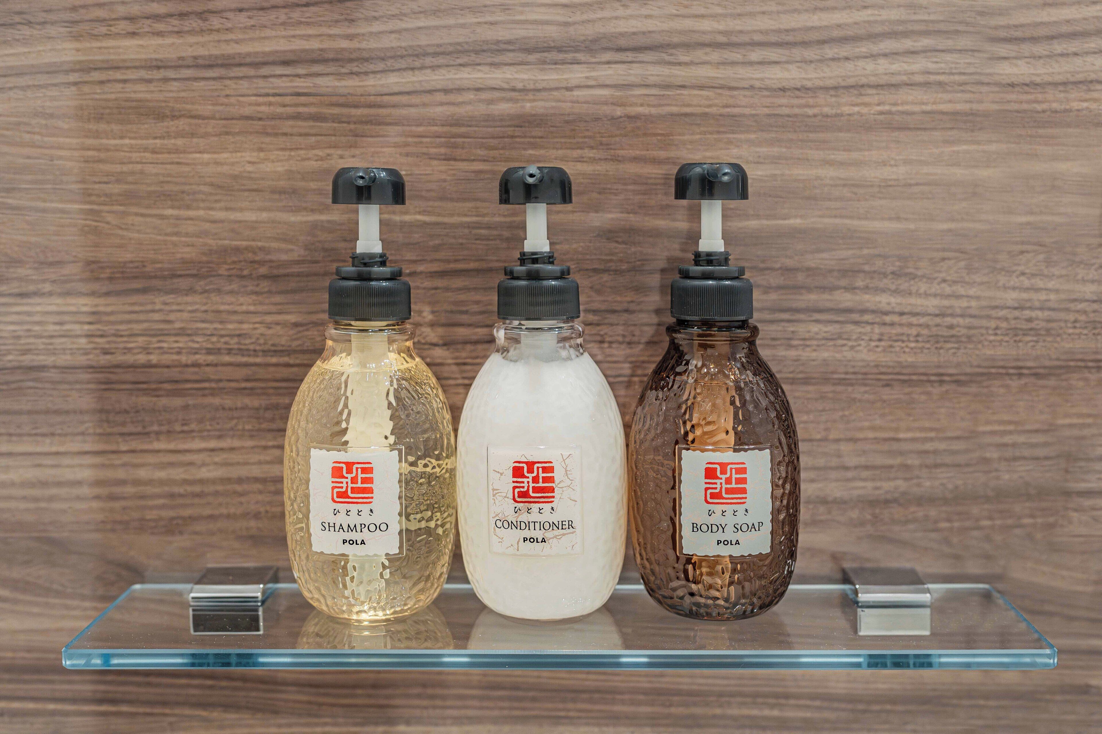 Three bottles of shampoo, conditioner, and body soap on a clear glass shelf against a wooden background.