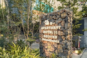 A sign for an apartment hotel with a stone wall and metal gate, surrounded by greenery. - Apartment Hotel 11 Shinsaibashi 9 (Osaka-shi)