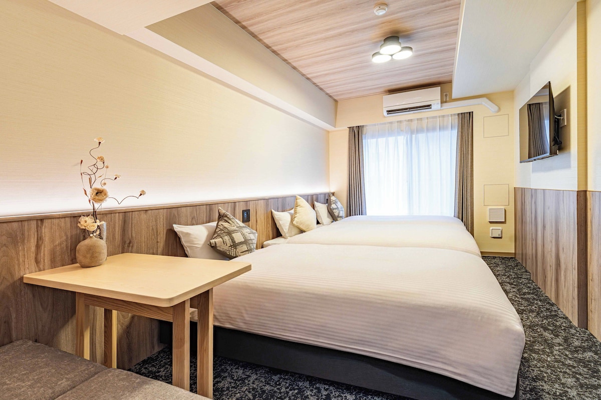A hotel room with a bed, bedside table, and a small desk with a vase of dried flowers.