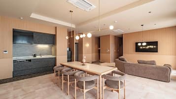 Una cocina moderna con horno empotrado, una zona de comedor con mesa de madera y sillas tapizadas en gris, y una sala de estar con sofá gris y televisor montado.