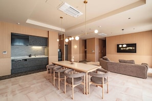 Una cocina moderna con horno empotrado, una zona de comedor con mesa de madera y sillas tapizadas en gris, y una sala de estar con sofá gris y televisor montado.