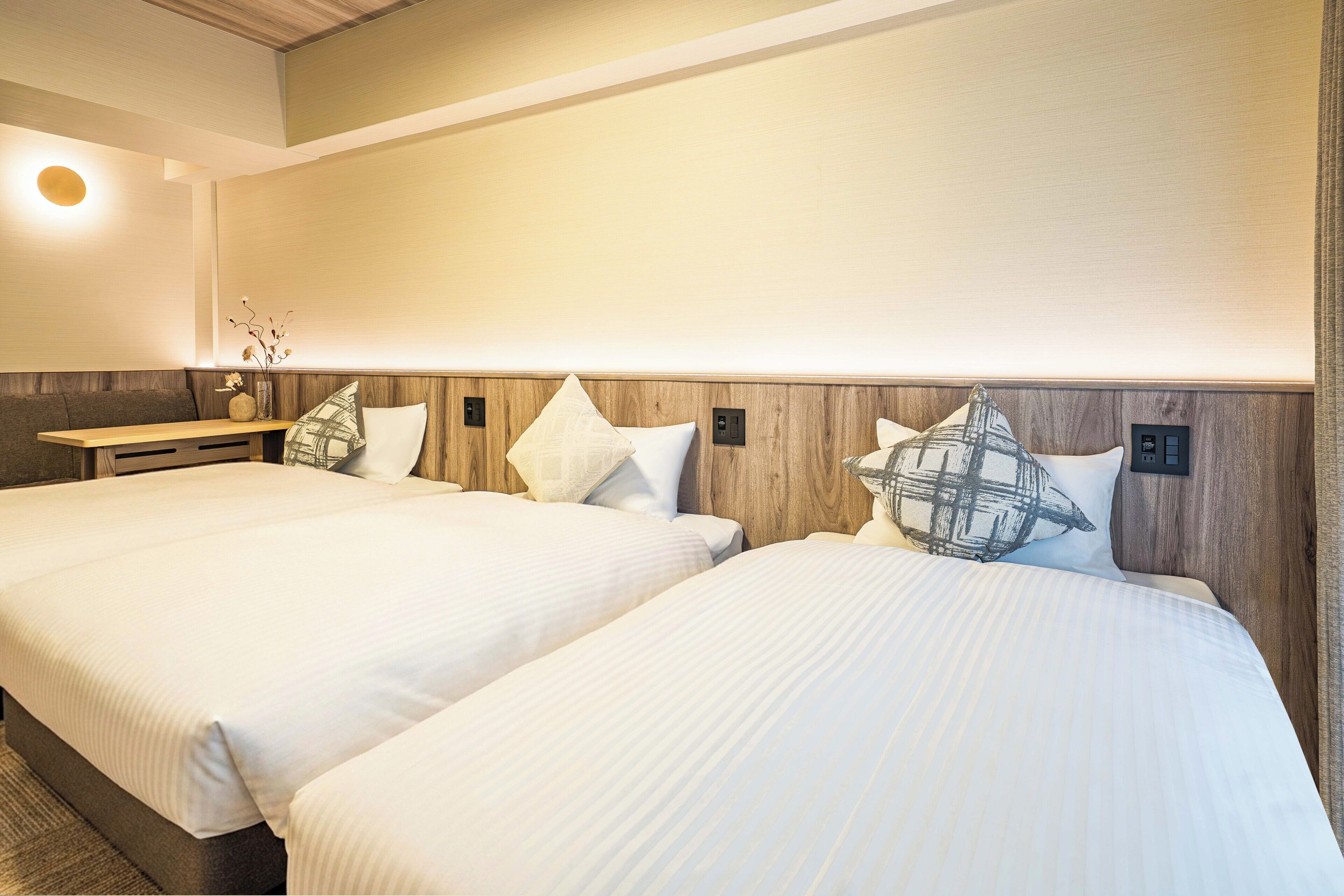 A hotel room with two beds, a wooden headboard, a bedside table, and a wall-mounted light fixture.