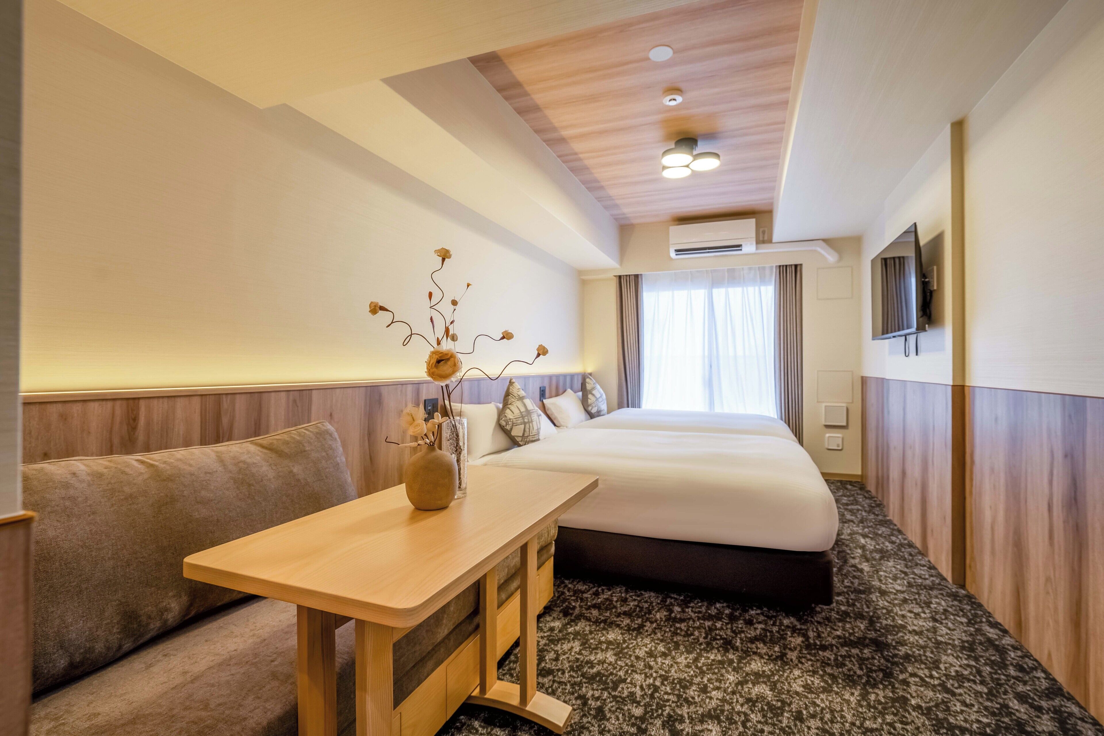 A hotel room with a bed, a small table with a vase, a sofa, and a television on the wall.
