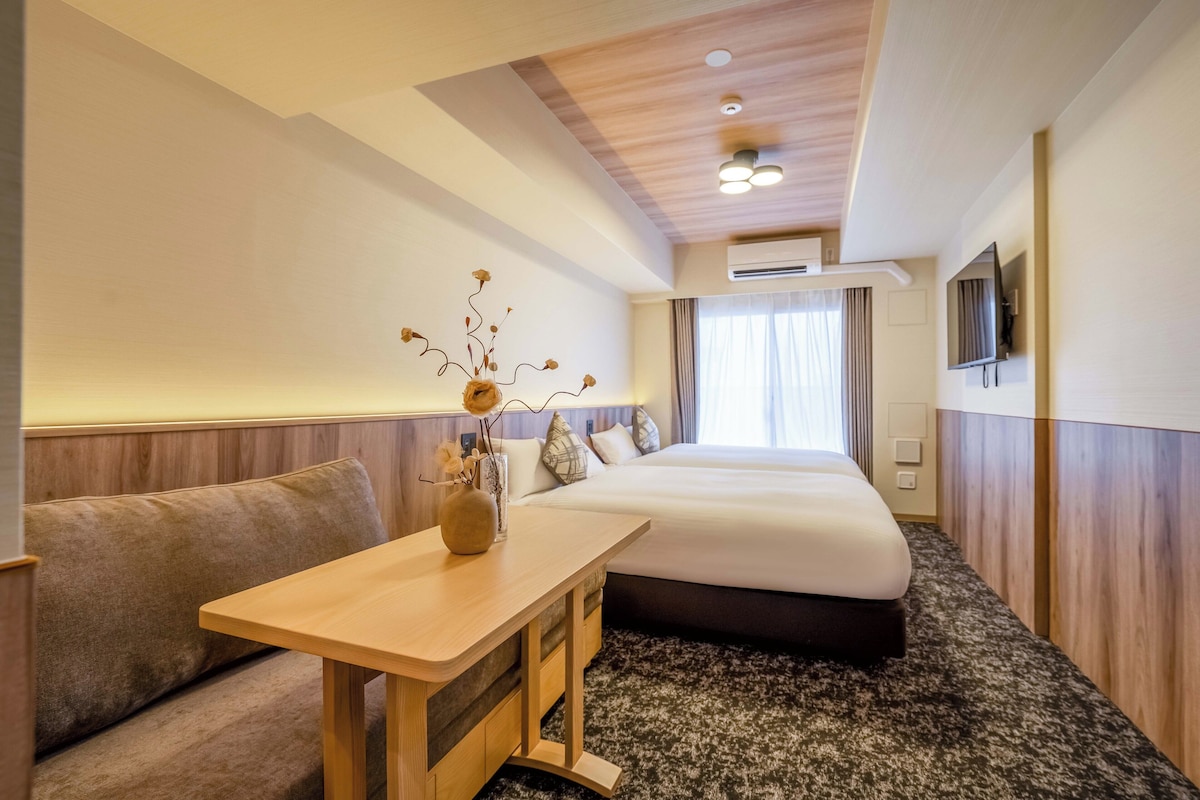 A hotel room with a bed, a small table with a vase, a sofa, and a television on the wall.