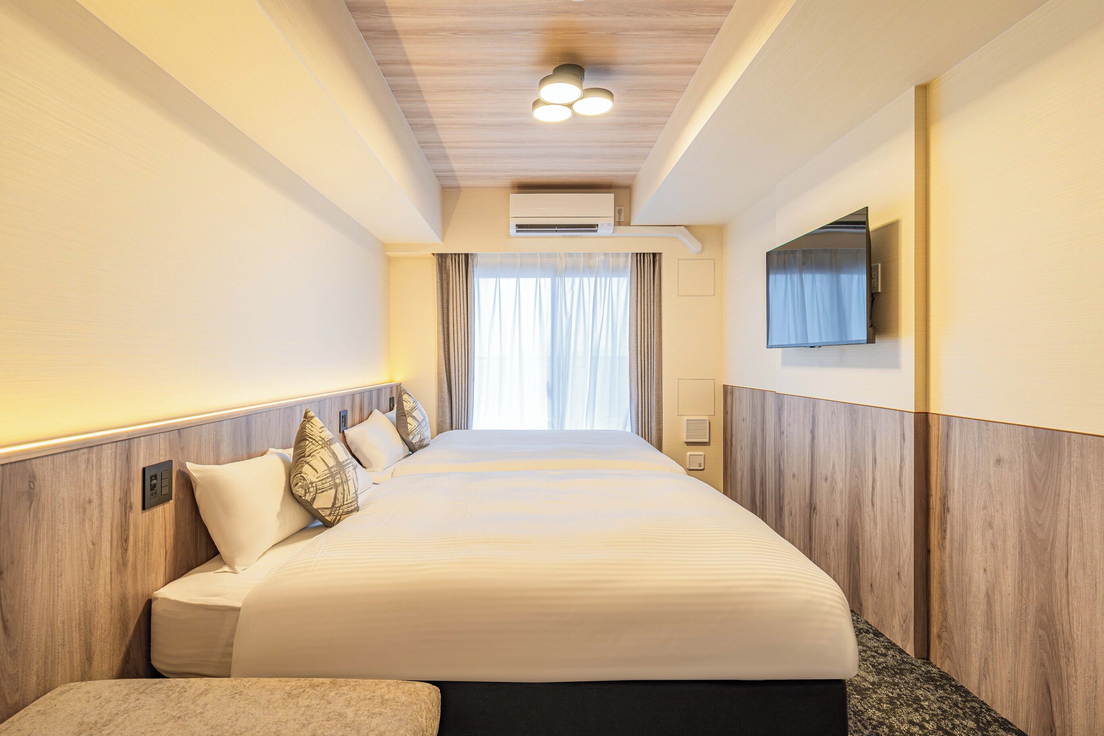 A hotel room with a large bed, a flat-screen TV mounted on the wall, and an air conditioning unit above the bed.