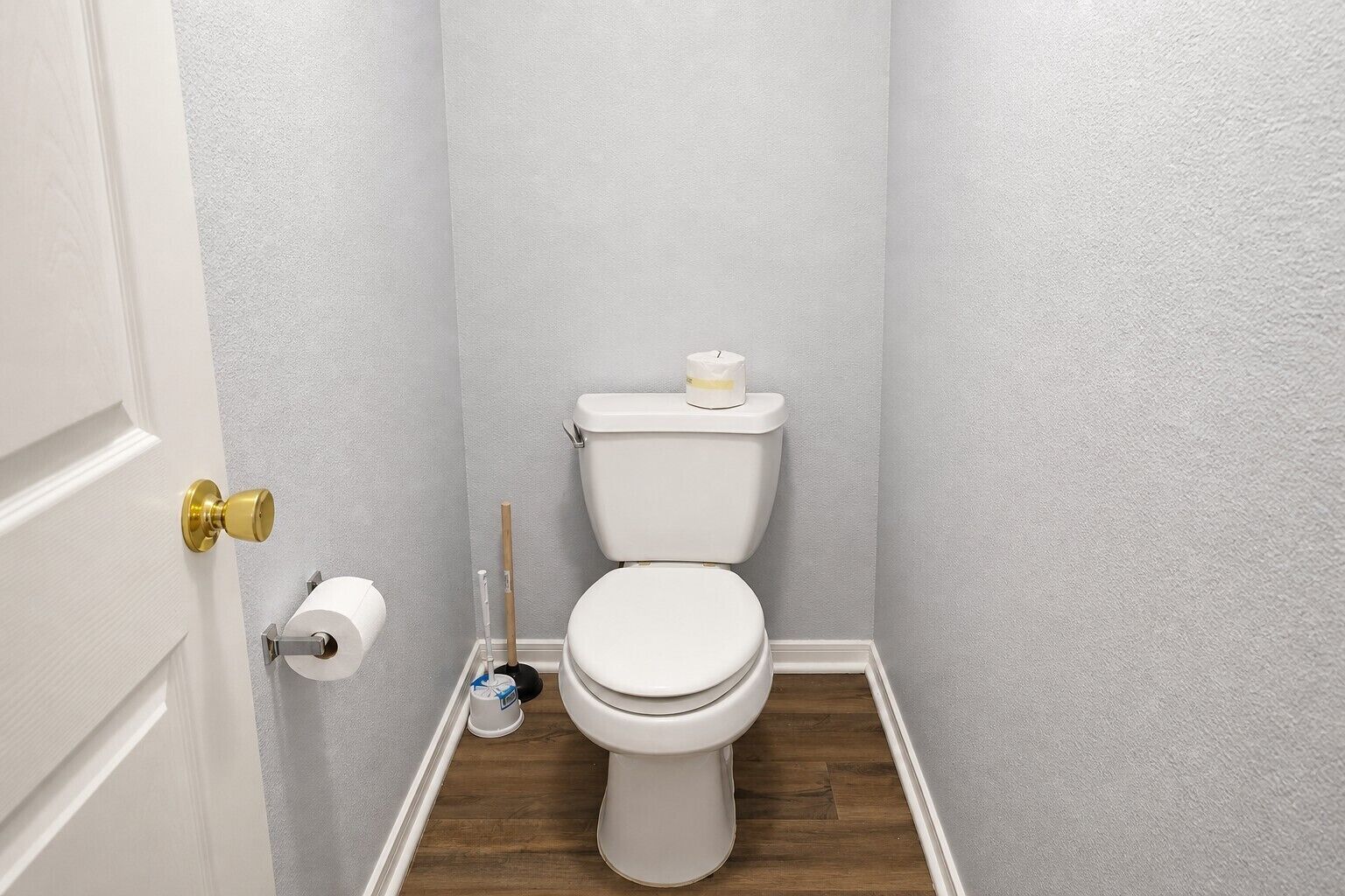 Ein kleines Badezimmer mit Toilette, Toilettenpapier, einem Besen und einem Mülleimer.