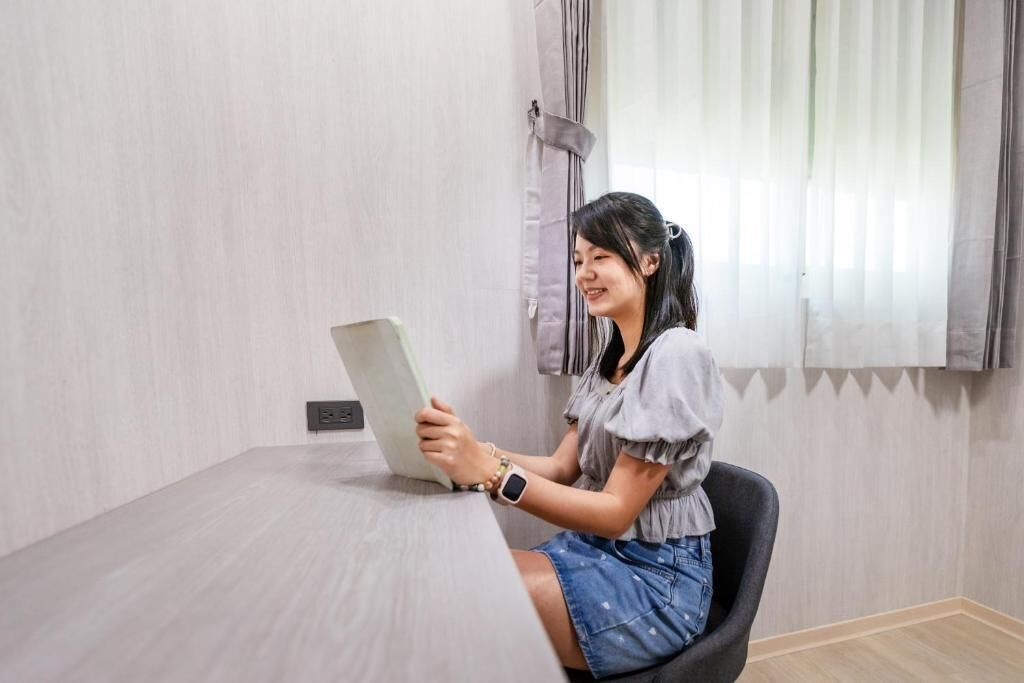 A person seated at a table in a room with a light-colored wall and a window with curtains.
