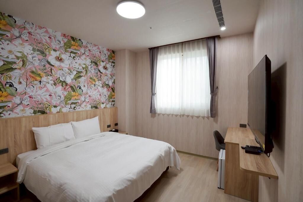 A hotel room with a bed, a desk with a chair, a flat-screen TV, and a window with curtains.