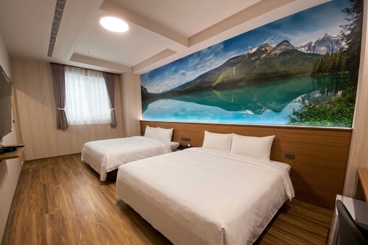 A hotel room with two beds, a large window, a painting of a mountain landscape, and a wooden floor.
