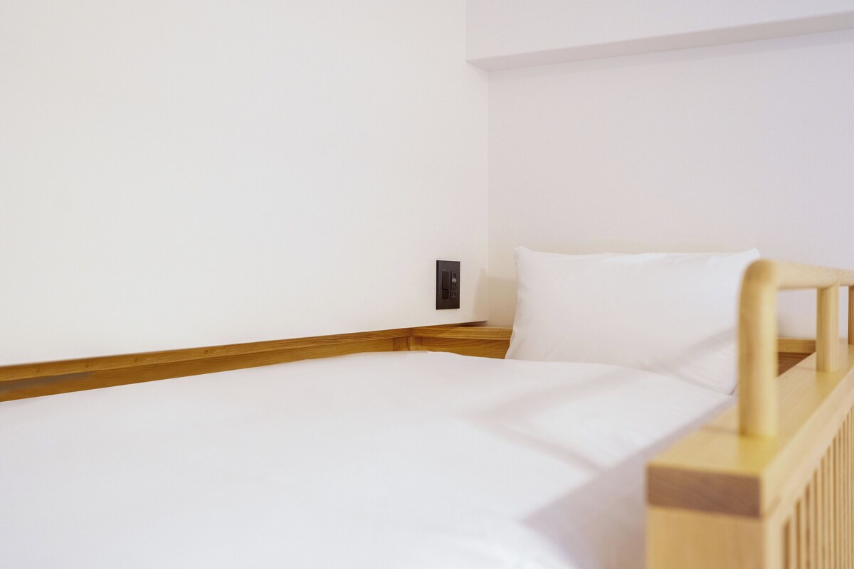 Ein Einzelbett mit Holz Kopfteil und weißer Bettwäsche in einem Zimmer mit schlichten weißen Wänden und Holzboden.