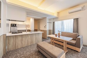 Ein modernes Hotelzimmer mit einer Küchenzeile, einem Sofa, einem kleinen Tisch und Blick auf die Stadt durch große Fenster.