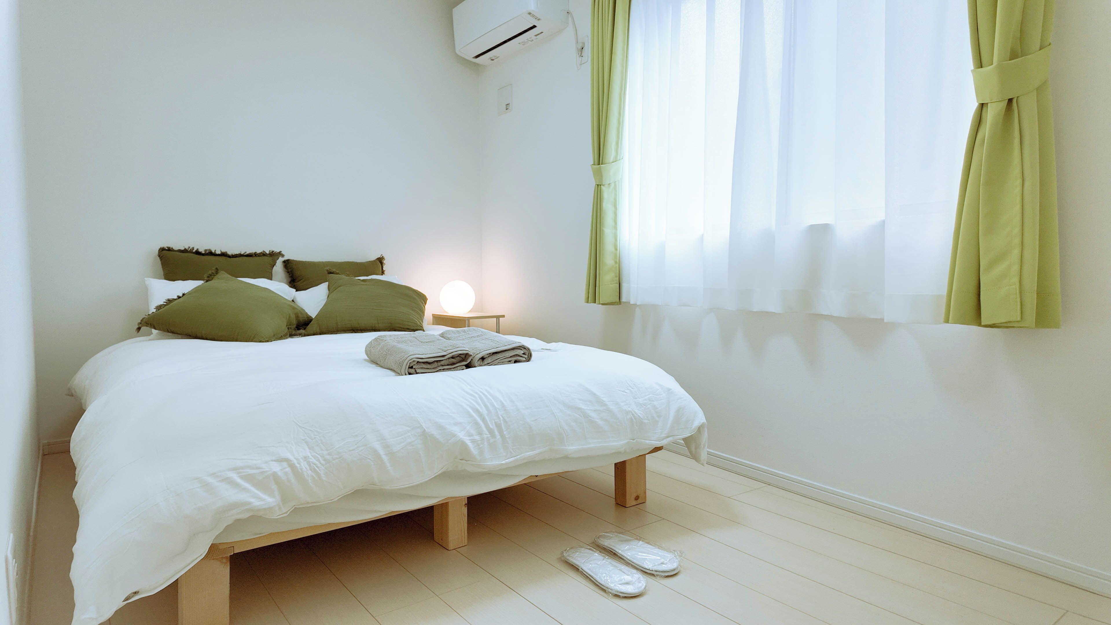 A neatly made bed with a white duvet and green pillows, a bedside lamp, and a window with sheer curtains.
