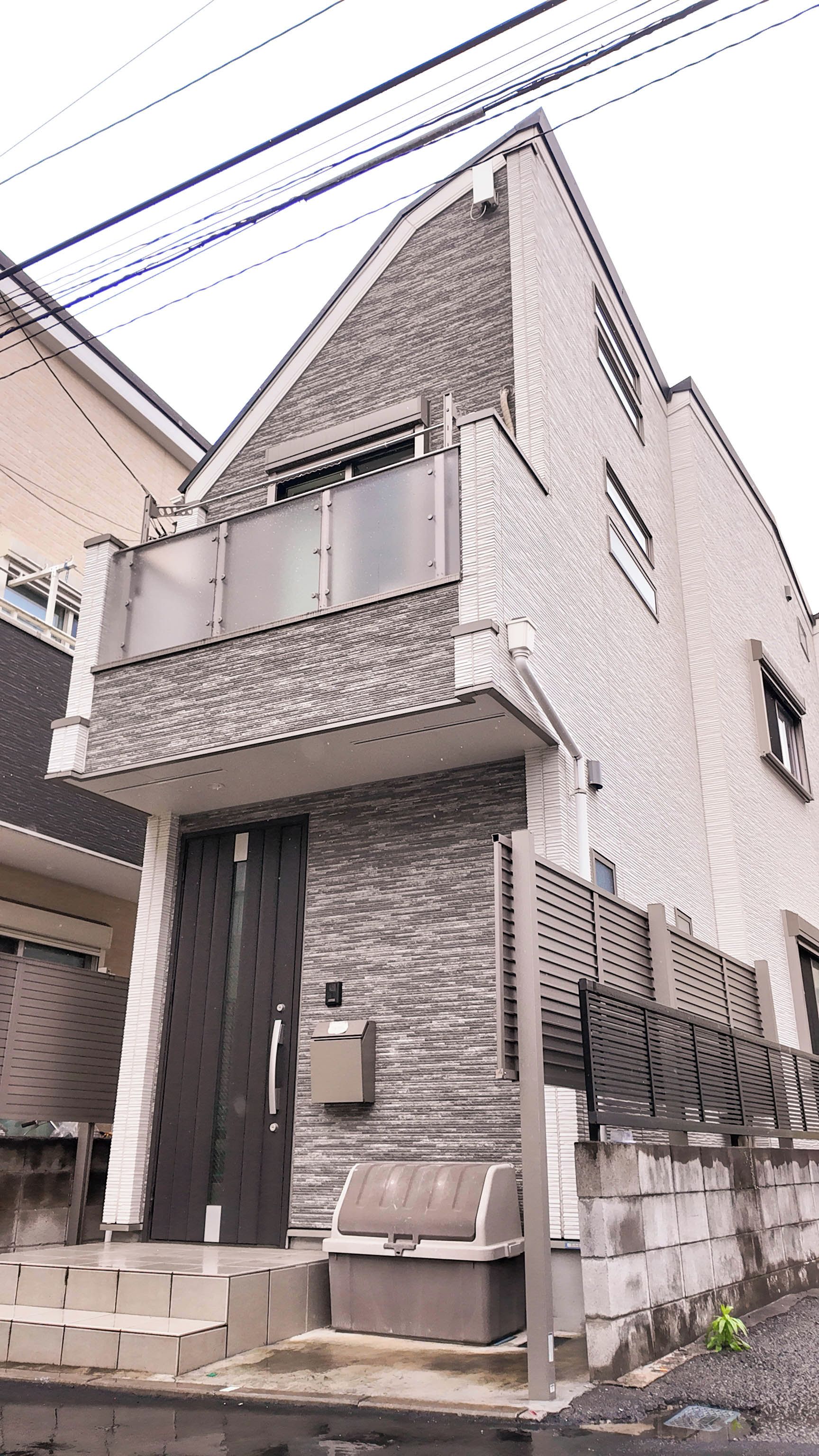 A two-story residential building with a dark front door, a small balcony, and a trash bin outside.