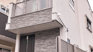 A two-story residential building with a dark front door, a small balcony, and a trash bin outside.
